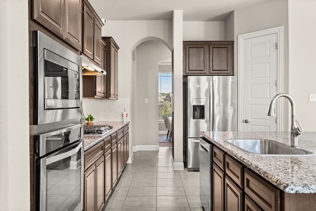 kitchen featuring arched walkways, under cabinet range hood, stainless steel appliances, a sink, and light stone countertops