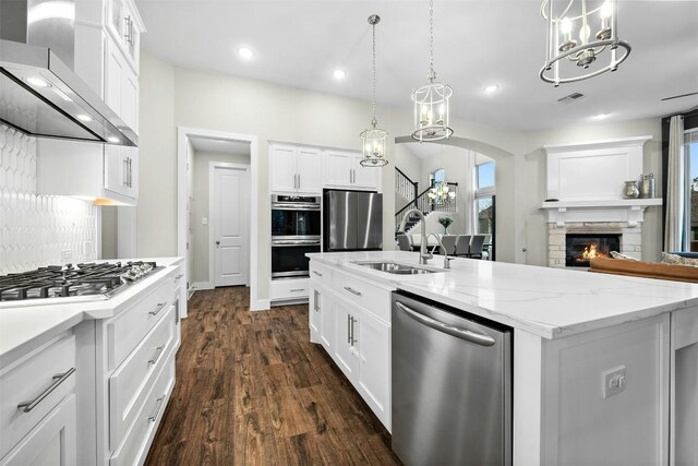 kitchen with white cabinetry, hanging light fixtures, appliances with stainless steel finishes, wall chimney range hood, and a center island with sink