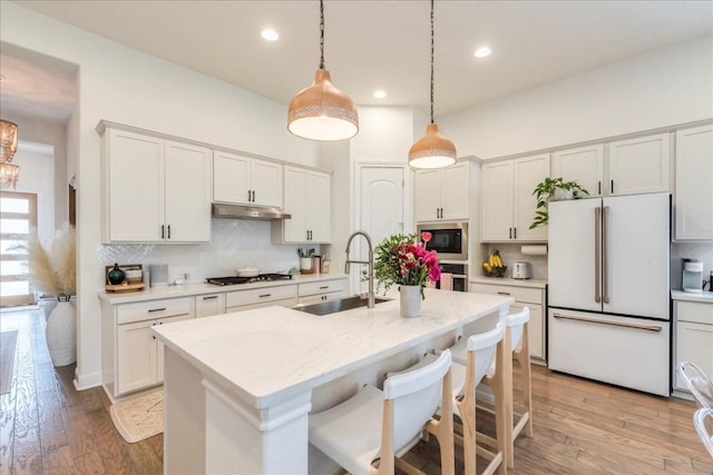 kitchen with a kitchen island with sink, under cabinet range hood, stainless steel appliances, a sink, and pendant lighting