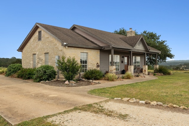 view of front facade featuring a porch, stone siding, roof with shingles, a chimney, and a front yard