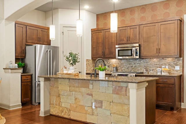 kitchen featuring decorative light fixtures, a center island with sink, appliances with stainless steel finishes, a sink, and dark stone counters