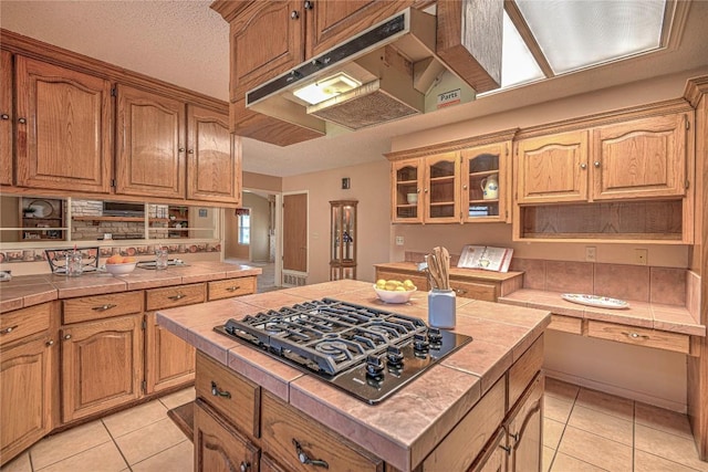kitchen with light tile patterned floors, tile counters, a kitchen island, glass insert cabinets, and stainless steel gas stovetop