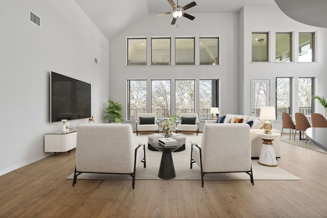 living area featuring baseboards, visible vents, a ceiling fan, a towering ceiling, and light wood-type flooring