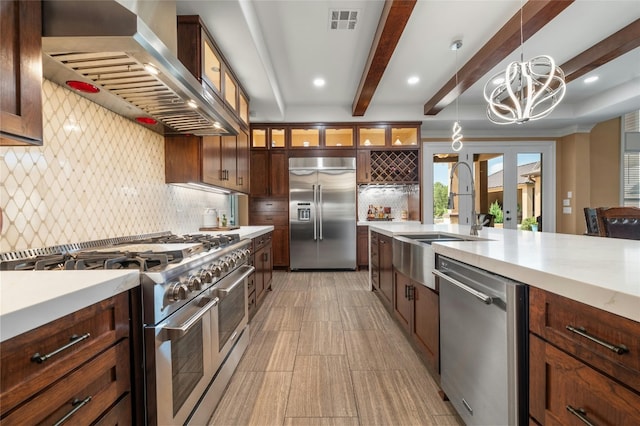 kitchen with premium appliances, light countertops, hanging light fixtures, wall chimney range hood, and glass insert cabinets