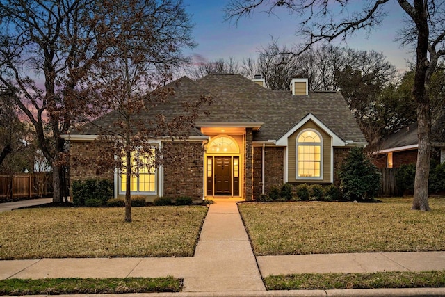 view of front of home with roof with shingles, brick siding, a chimney, a lawn, and fence