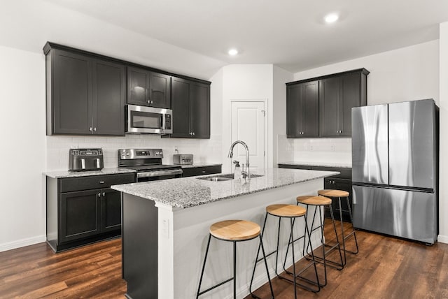 kitchen featuring dark wood-style flooring, a center island with sink, stainless steel appliances, a sink, and light stone countertops