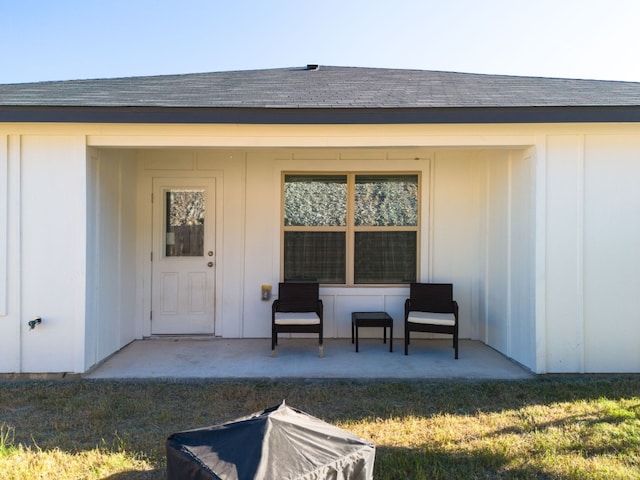 view of exterior entry featuring a yard, board and batten siding, a patio area, and roof with shingles