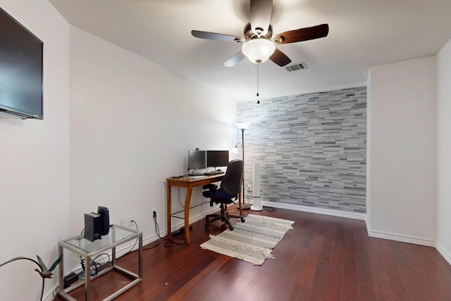 office area with an accent wall, visible vents, baseboards, a ceiling fan, and dark wood finished floors