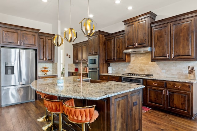 kitchen featuring under cabinet range hood, dark wood-style flooring, appliances with stainless steel finishes, decorative backsplash, and an island with sink