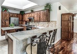 kitchen featuring under cabinet range hood, a peninsula, appliances with stainless steel finishes, backsplash, and a kitchen bar