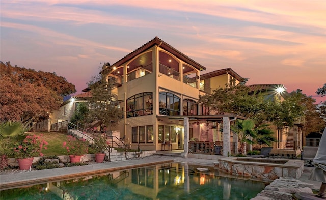 back of house at dusk featuring a balcony, stairs, a patio area, a pool with connected hot tub, and stucco siding