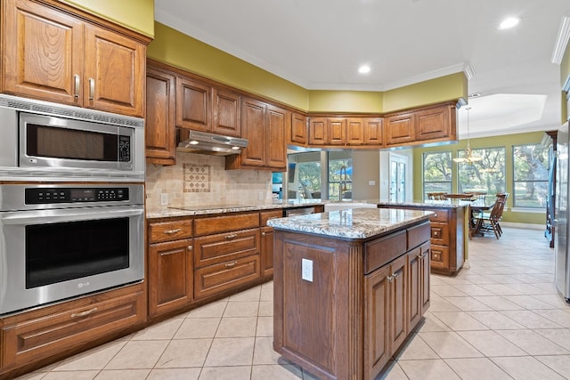 kitchen with a center island, brown cabinets, stainless steel appliances, light stone countertops, and under cabinet range hood