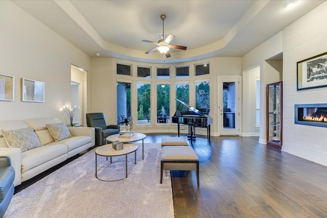 living area with dark wood-style flooring, a raised ceiling, a ceiling fan, a tile fireplace, and baseboards