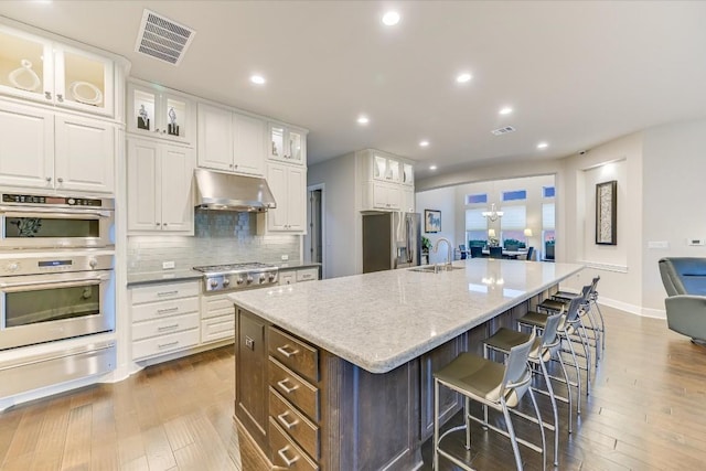 kitchen featuring visible vents, glass insert cabinets, a kitchen island with sink, white cabinets, and under cabinet range hood