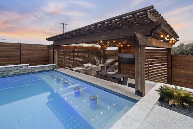 pool at dusk featuring fence, an outdoor kitchen, a pergola, and area for grilling