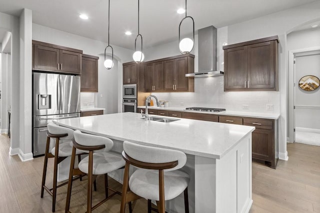 kitchen with a sink, hanging light fixtures, appliances with stainless steel finishes, wall chimney exhaust hood, and an island with sink