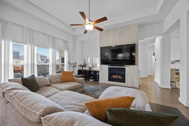 living room featuring ceiling fan, a large fireplace, baseboards, dark wood-style floors, and a tray ceiling