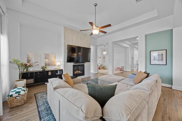 living room featuring a raised ceiling, light wood-style flooring, a ceiling fan, a large fireplace, and baseboards