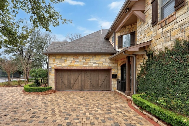 exterior space with decorative driveway, stone siding, roof with shingles, and a garage