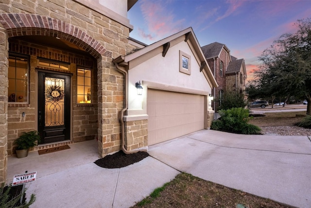 exterior entry at dusk with a garage, stone siding, concrete driveway, and stucco siding