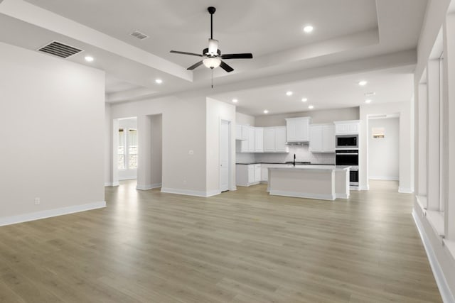 unfurnished living room featuring light wood-type flooring, ceiling fan, visible vents, and a raised ceiling