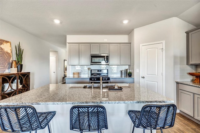 kitchen featuring gray cabinets, light wood-style flooring, appliances with stainless steel finishes, a kitchen island with sink, and light stone countertops