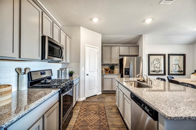 kitchen with light stone counters, dark wood-type flooring, a sink, appliances with stainless steel finishes, and a center island with sink