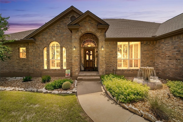 entrance to property with a shingled roof, a lawn, and brick siding