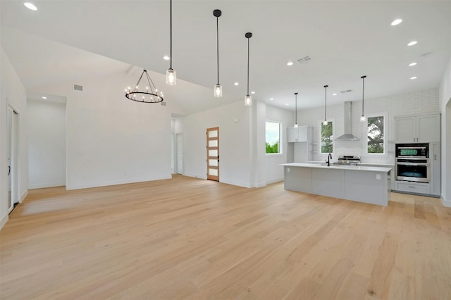 kitchen with open floor plan, light countertops, wall chimney range hood, an island with sink, and pendant lighting