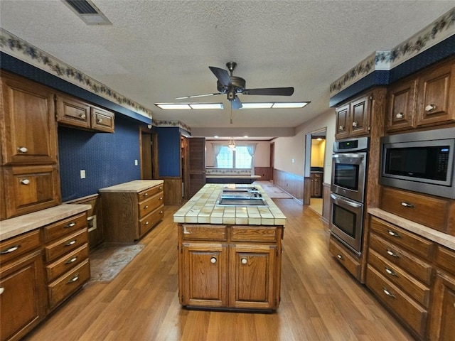 kitchen with wainscoting, tile countertops, appliances with stainless steel finishes, a center island, and light wood-style floors