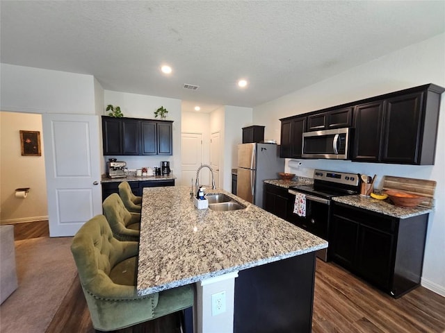 kitchen featuring an island with sink, light stone counters, stainless steel appliances, a kitchen bar, and a sink