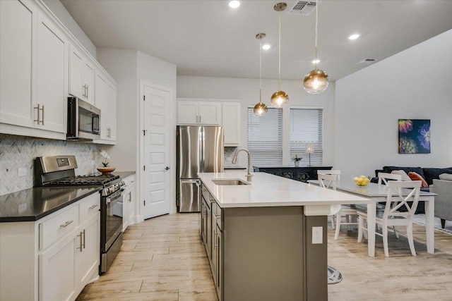 kitchen with white cabinets, dark countertops, appliances with stainless steel finishes, hanging light fixtures, and a kitchen island with sink