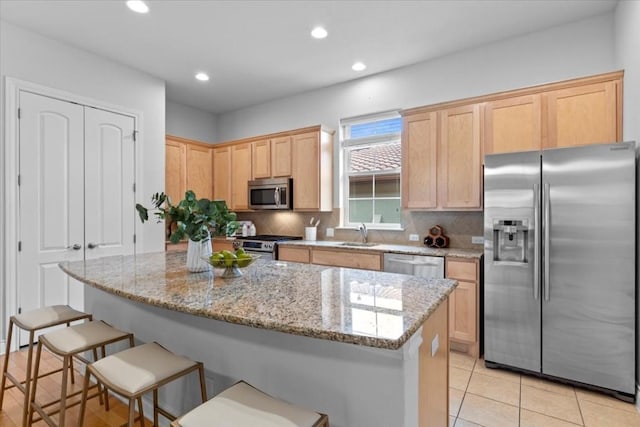 kitchen with light stone counters, stainless steel appliances, a kitchen island, light brown cabinetry, and tasteful backsplash