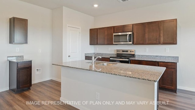 kitchen featuring a kitchen island with sink, stainless steel appliances, dark wood-style flooring, a sink, and light stone countertops