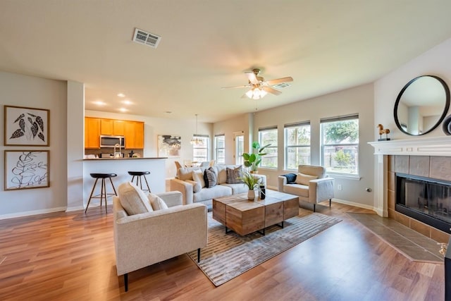 living area with ceiling fan, a tile fireplace, visible vents, baseboards, and light wood-type flooring