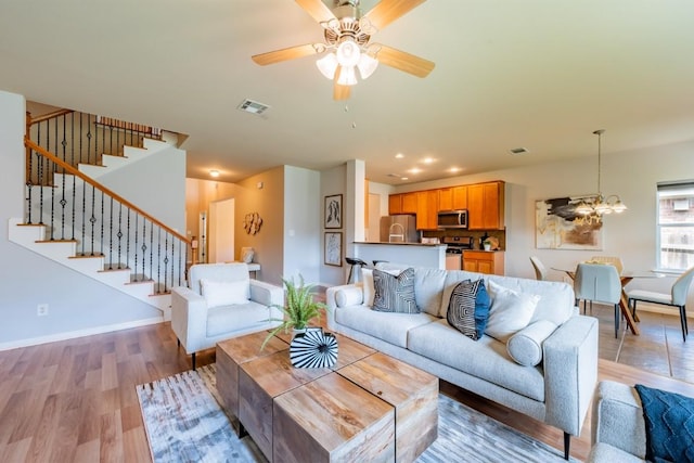 living room featuring visible vents, light wood-style flooring, stairway, baseboards, and ceiling fan with notable chandelier