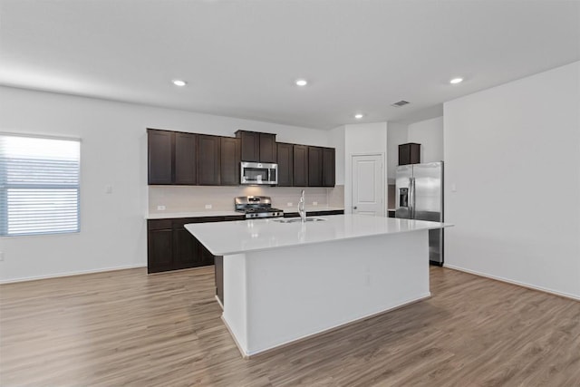 kitchen with dark brown cabinetry, stainless steel appliances, a sink, light countertops, and an island with sink