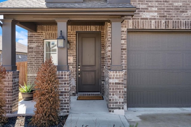 view of exterior entry with an attached garage, a shingled roof, and brick siding