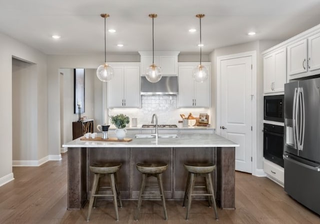 kitchen with under cabinet range hood, light countertops, black appliances, a center island with sink, and pendant lighting