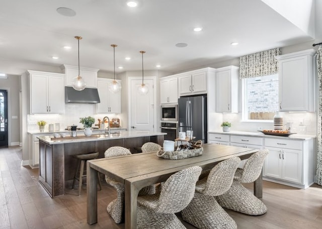 kitchen with pendant lighting, light countertops, freestanding refrigerator, a kitchen island with sink, and under cabinet range hood