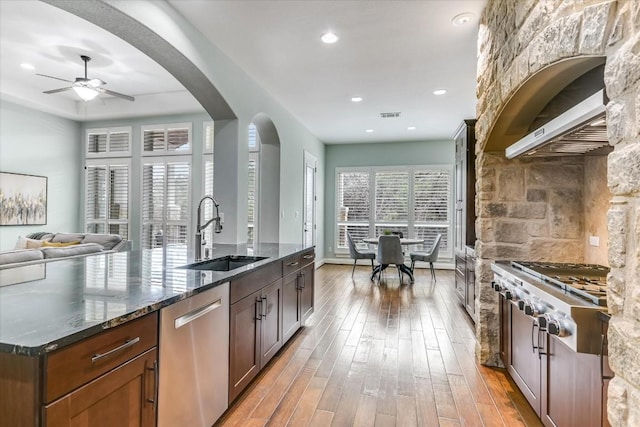 kitchen featuring arched walkways, dark stone counters, a kitchen island with sink, stainless steel appliances, and a sink