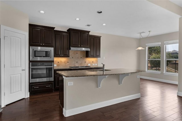 201 Seminole Canyon Dr, Georgetown, TX 78628 - kitchen featuring stainless steel appliances, under cabinet range hood, dark stone counters, a kitchen island with sink, and backsplash