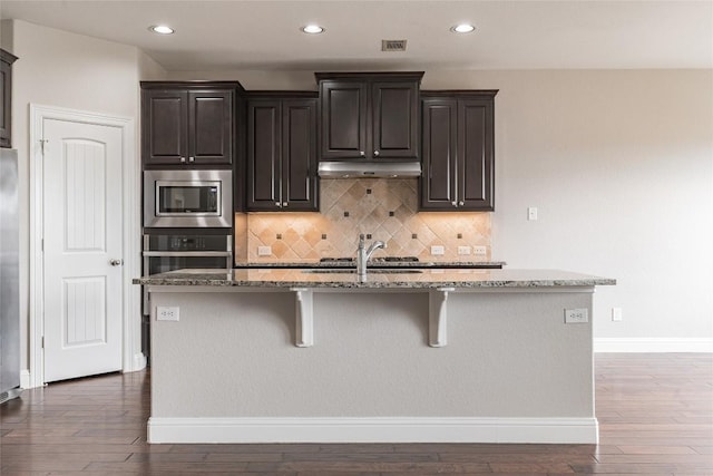 kitchen featuring a breakfast bar area, stone countertops, a kitchen island with sink, under cabinet range hood, and stainless steel appliances