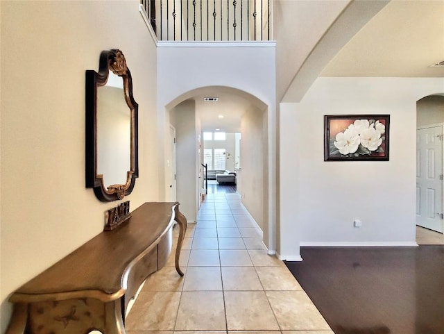 hall featuring arched walkways, light tile patterned floors, visible vents, a towering ceiling, and baseboards