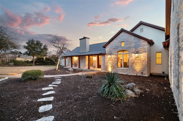 back of property featuring driveway, stone siding, a chimney, and metal roof