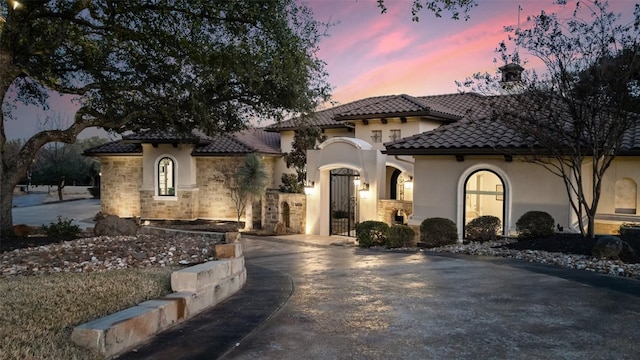 mediterranean / spanish house featuring concrete driveway, stone siding, a tile roof, a gate, and stucco siding