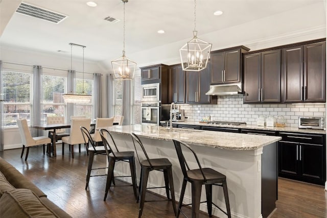 kitchen featuring a breakfast bar area, visible vents, hanging light fixtures, an island with sink, and under cabinet range hood