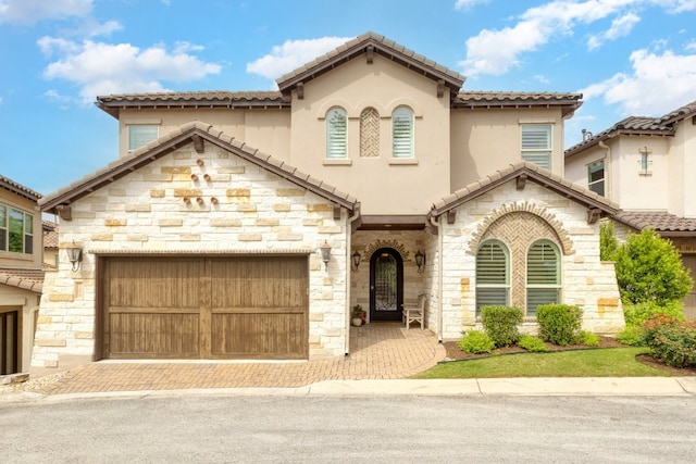 mediterranean / spanish-style house with decorative driveway, a tile roof, stucco siding, an attached garage, and stone siding