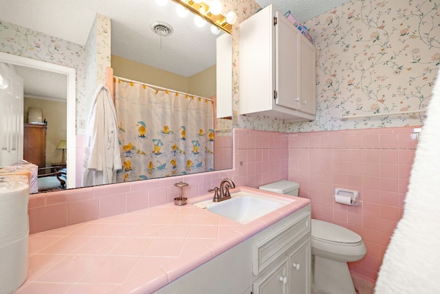 full bathroom with a textured ceiling, vanity, tile walls, wainscoting, and wallpapered walls