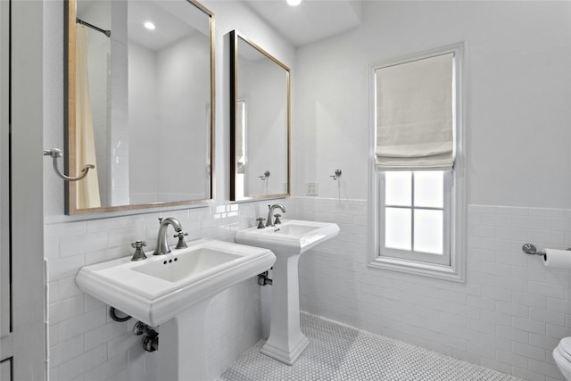 full bath with two sinks, toilet, a wainscoted wall, tile patterned flooring, and tile walls